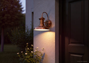 The Urban Ambiance UHP1023 Luxe Industrial Outdoor Wall Light from the Palermo Collection, featuring a solid copper finish and measuring 10.75″ high by 10″ wide, is mounted beside a dark door, casting its vintage-inspired glow over white flowers and part of a stucco wall at dusk.