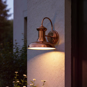The UHP1023 Luxe Industrial Outdoor Wall Light from the Palermo Collection by Urban Ambiance, featuring a solid copper finish and measuring 10.75″ high by 10″ wide, is mounted on a stucco wall. Reminiscent of vintage exterior lighting, this stylish fixture casts a warm glow at dusk, with greenery and white flowers in the foreground.