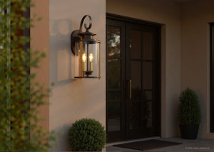 The Urban Ambiance UHP1120 Rustic Outdoor Wall Light from the Plymouth Collection, featuring a 19.25″H x 8″W silhouette and an Olde Bronze finish, is installed beside the front door; two potted shrubs and a doormat on the porch enhance the inviting entryway.