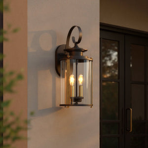 The UHP1120 Rustic Outdoor Wall Light from Urban Ambiance’s Plymouth Collection, measuring 19.25″ high by 8″ wide and finished in Olde Bronze, features two visible bulbs and clear glass panels. Mounted on a beige wall near glass doors, it casts a warm glow at dusk, seamlessly blending rustic modern style with classic elegance.