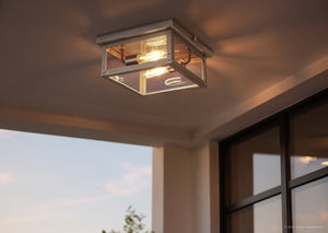 The Urban Ambiance UHP1133 Modern Farmhouse Outdoor Ceiling light from the Darwin Collection, featuring a sleek stainless steel finish and measuring 5-1/2″ high by 12-3/8″ wide, is mounted on an outdoor porch ceiling at dusk, its exposed bulbs casting a warm glow.