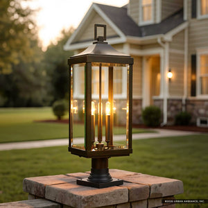 The Urban Ambiance UHP1190 Colonial Outdoor Post / Pier Light from the Calderdale Collection, featuring an Olde Bronze finish and beveled glass panels with four visible bulbs, is mounted on a stone pillar outside a house, casting a warm glow over the green lawn in the evening.
