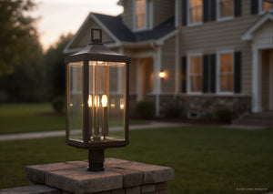 The Urban Ambiance UHP1190 Colonial Outdoor Post / Pier Light from the Calderdale Collection, measuring 26-7/8″ x 9-1/2″ and finished in Olde Bronze, is mounted on a brick post. The four-bulb lantern glows at dusk with a beveled glass design, set against the backdrop of a suburban house and yard.