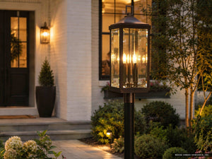 An Urban Ambiance UHP1190 Colonial Outdoor Post / Pier Light from the Calderdale Collection, featuring an Olde Bronze finish and beveled glass, stands illuminated in a landscaped garden near the entrance of a house with white brick walls and a black door.