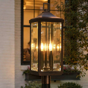 A lit UHP1190 Colonial Outdoor Post / Pier Light from the Calderdale Collection by Urban Ambiance, measuring 26-7/8″ x 9-1/2″ with a striking Olde Bronze finish and clear glass panels, is mounted on a post; a window and plants are visible in the background.