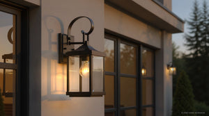 The UHP1192 Colonial Outdoor Wall Light from Urban Ambiance’s Calderdale Collection features an Olde Bronze finish, clear beveled glass, and a visible bulb. Measuring 15-1/8″ x 5-1/2″, this sconce is shown mounted on a beige exterior wall at sunset with trees in the background, bringing timeless Colonial charm to your home’s facade.