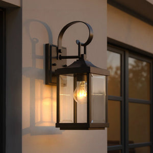 An Urban Ambiance UHP1192 Colonial Outdoor Wall Light from the Calderdale Collection, featuring an Olde Bronze finish and measuring 15-1/8″ by 5-1/2″, is mounted beside a window on a beige wall at dusk, its clear beveled glass panels and black metal frame showcasing a softly glowing bulb.
