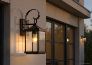 An Urban Ambiance UHP1192 Colonial Outdoor Wall Light from the Calderdale Collection, measuring 15-1/8″ x 5-1/2″ and featuring an Olde Bronze finish with clear beveled glass and a visible bulb, is mounted on the exterior of a modern home near the windows.