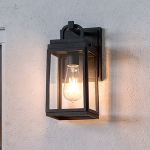 The UHP1385 Farmhouse Outdoor Wall Sconce from Urban Ambiance's Macon Collection features a marine-grade, powder-coated midnight black finish with metal and glass construction, mounted on a light-textured wall and illuminated by an exposed bulb.