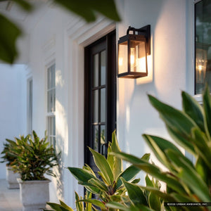 The Urban Ambiance UHP1385 Farmhouse Outdoor Wall Sconce, featuring a midnight black marine-grade finish from the Macon Collection, enhances a white home’s porch, complementing potted green plants.