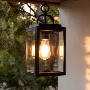 The Urban Ambiance UHP1388 Macon Farmhouse Outdoor Pendant (15.375''H x 7''W) in midnight black features marine-grade metal and glass, hanging from the ceiling with a visible bulb, set against a backdrop of trees.
