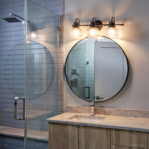 Modern bathroom with a round mirror, the Urban Ambiance UHP2044 Luxe Industrial Bath Fixture from the Harlow Collection in a Fashion Bronze finish featuring prismatic glass, wood vanity, stone countertop, and glass shower with tiled wall.
