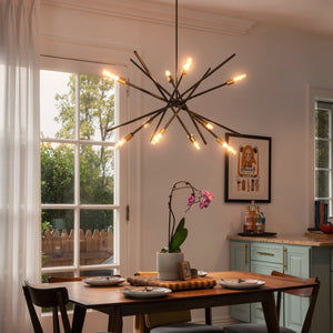 Modern dining room with a wooden table set for four, a potted orchid centerpiece, and sunlight streaming through glass doors. The space is illuminated by the Urban Ambiance UHP2070 Modern Industrial Chandelier from the Miami Collection, featuring a striking Olde Bronze finish and 19-5/8″ x 42-3/8″ dimensions that add a touch of modern vintage style to this inviting setting.