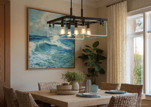 Dining area featuring woven chairs and a wooden table set for four, illuminated by the Urban Ambiance Bristol Collection UHP2444 Modern Farmhouse Square Chandelier in an Olde Bronze finish, with a large ocean painting, potted plant, and a window dressed in cream curtains.