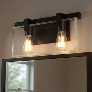 The Urban Ambiance UHP2452 Modern Farmhouse Modern Bath Fixture from the Bristol Collection, featuring an olde bronze finish and measuring 8-3/8″ high by 14-7/8″ wide, is mounted above a rectangular mirror. Its two exposed bulbs and sleek metal-and-glass design evoke a contemporary farmhouse look while reflecting a window view.