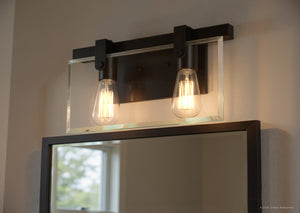 Above a rectangular mirror, the Urban Ambiance UHP2452 Modern Farmhouse Modern Bath Fixture from the Bristol Collection (8-3/8″H x 14-7/8″W, Olde Bronze Finish) with two exposed bulbs casts light, reflecting part of a window and a white wall.
