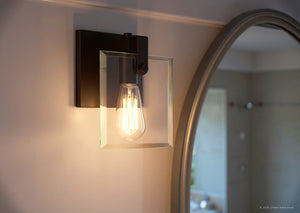 The Urban Ambiance UHP2454 Modern Farmhouse Bath/Wall Light from the Bristol Collection, featuring an exposed bulb and a square beveled glass plate in Olde Bronze finish (8-3/8″H x 7″W), is shown illuminated next to a round mirror in a softly lit room.