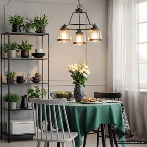 A dining room featuring a green tablecloth on the table, black chairs, and a black shelf adorned with plants and dishes, is illuminated by the Urban Ambiance UHP2533 Luxe Industrial Chandelier from the Nottingham Collection. This artisan-style chandelier showcases glass globes in an Olde Bronze finish and hangs above the table near a window with sheer curtains.