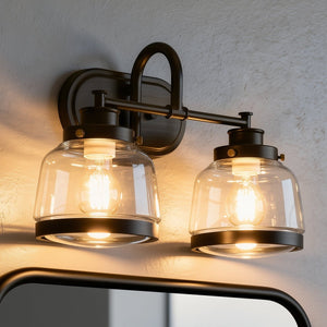 The Urban Ambiance UHP2537 Luxe Industrial Bathroom Vanity Light from the Nottingham Collection, measuring 11.25″ high by 16.25″ wide and finished in Olde Bronze, features two clear glass shades with exposed bulbs, elegantly mounted above a mirror on a white wall.