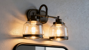 The UHP2537 Luxe Industrial Bathroom Vanity Light from the Nottingham Collection by Urban Ambiance is a wall-mounted fixture installed above a mirror on a textured wall. Featuring an Olde Bronze finish and vintage metal fittings, this 11.25″H x 16.25″W light offers two glass shades with exposed bulbs that emit warm illumination.