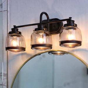 The Urban Ambiance UHP2539 Luxe Industrial Bathroom Vanity Light from the Nottingham Collection, featuring three clear glass shades and an Olde Bronze finish (11.25″H x 26″W), is wall-mounted above a round bathroom mirror and beautifully illuminates a textured wall.