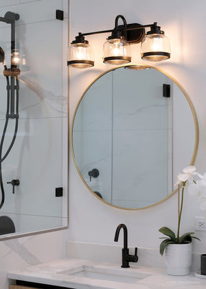 Modern bathroom with a round mirror, black faucet, bath fixture in olde bronze, the Urban Ambiance UHP2539 Luxe Industrial Bathroom Vanity Light from the Nottingham Collection (11.25″H x 26″W) in olde bronze finish, three schoolhouse globe lights, glass shower, white marble countertop, and a potted orchid.