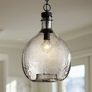 The UHP2770 Modern Farmhouse Pendant from the Hobart Collection by Urban Ambiance features a clear, hammered glass shade that hangs elegantly from the ceiling, accentuating a visible light bulb within. Its charcoal finish and artisan craftsmanship are subtly highlighted by softly lit, blurred windows in the background, enhancing the pendant’s refined modern farmhouse appeal.