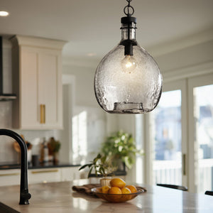 The Urban Ambiance UHP2770 Modern Farmhouse Pendant from the Hobart Collection, featuring a textured glass shade and visible bulb in a charcoal finish, hangs above a marble kitchen island with a bowl of lemons; in the background, white cabinets enhance the bright, modern ambiance.