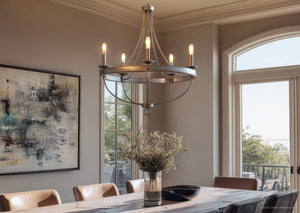 A modern dining room features a large window, abstract wall art, and a wooden table adorned with a vase of white flowers, illuminated by the Urban Ambiance UHP2900 Farmhouse Chandelier from the Adelaide Collection in a galvanized steel finish (30″ x 26″).