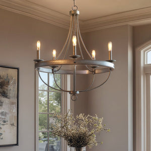 The Urban Ambiance UHP2900 Farmhouse Chandelier from the Adelaide Collection, featuring a 30″ x 26″ galvanized steel finish and six exposed bulbs, hangs from the ceiling of a room with large windows, a framed painting, and a vase of flowers on the table below.