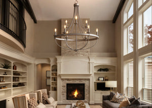 A spacious living room featuring the Urban Ambiance UHP2902 XL Farmhouse Chandelier from the Adelaide Collection, showcasing a striking open cage design in a galvanized steel finish, complemented by a brick fireplace, built-in shelves, comfortable sofas, a TV, and tall windows that offer scenic outdoor views.