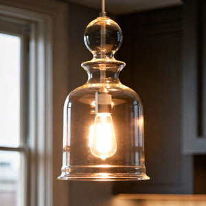 A glowing bulb is suspended inside the hand-blown glass shade of the Urban Ambiance UHP3043 Modern Farmhouse Pendant Light from the Dundee Collection, its 16-1/2″ x 9″ silhouette and brushed nickel finish lending a touch of modern elegance as it hangs beside a window.