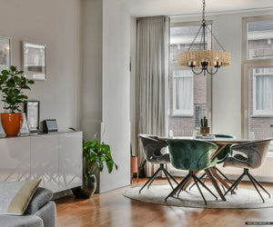 A contemporary dining room featuring a round table and four chairs is complemented by the Urban Ambiance UHP4781 Casual Indoor Chandelier from the Bexley Collection. This chandelier, with its vintage-style bulbs and matte black finish, is 21-1/2'' high and 20'' wide x 20'' deep. Large windows emphasize the indoor plants and artwork adorning the space.