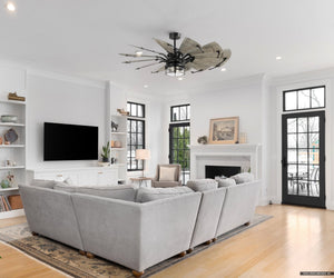 A spacious living room showcases a large sectional sofa, wall-mounted TV, and fireplace. Accented by the Urban Ambiance UHP9478 Tuscan Indoor Ceiling Fan in matte black, its tall windows and glass doors lead to a patio.