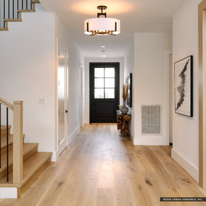 A bright hallway features light wood floors, white walls, a black door at the end, and a staircase on the left. Overhead, the Urban Ambiance Oakmont Collection UKC20373 Transitional Indoor Ceiling Light (14.5''H x 20''W x 20''Dep) with a sleek black finish adds a modern, inviting touch to the space.