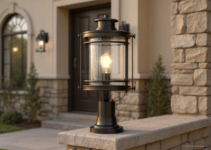 An Urban Ambiance UQL1010 Mid-Century Modern Outdoor Post Light from the Cannes Collection, featuring a black silk finish and seeded glass, is illuminated and placed on a stone ledge in front of a house with stone and stucco exterior walls.
