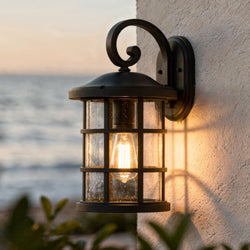 A lit Urban Ambiance UQL1044 Vienna Collection Craftsman outdoor wall light in natural black, 17.75"H x 10"W, with marine-grade construction is mounted on a white textured wall against an ocean sunset backdrop.