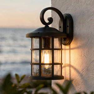 A lit Urban Ambiance UQL1044 Vienna Collection Craftsman outdoor wall light in natural black, 17.75"H x 10"W, with marine-grade construction is mounted on a white textured wall against an ocean sunset backdrop.