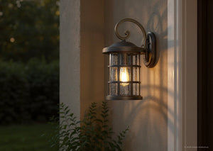 A UQL1045 Craftsman Outdoor Wall Light from Urban Ambiance’s Vienna Collection, featuring a Parisian Bronze finish and marine-grade construction, is mounted on a beige wall near a door. Its seeded glass softly illuminates the entrance, while greenery appears in the background.