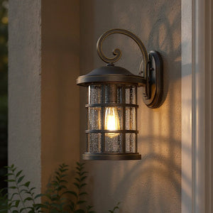 The Urban Ambiance UQL1045 Craftsman Outdoor Wall Light from the Vienna Collection, featuring a Parisian Bronze finish and marine-grade construction, is mounted on a stucco wall. Measuring 17.75 inches high by 10 inches wide, this fixture includes a clear seeded glass shade and is beautifully illuminated by a visible light bulb.