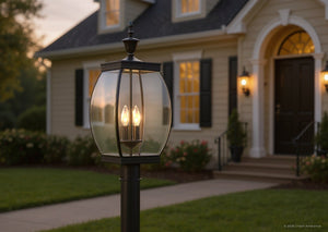 The UQL1174 Colonial Outdoor Post Light from the Manchester Collection by Urban Ambiance, featuring a 22-inch height, 9-inch width, and Medieval Bronze finish, stands on a lawn in front of a beige house with illuminated windows, a black door, and flower beds bordering the walkway at dusk.