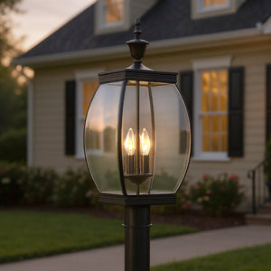 The Urban Ambiance UQL1174 Colonial Outdoor Post Light from the Manchester Collection, featuring a medieval bronze finish and measuring 22 inches high by 9 inches wide, stands illuminated with its glass panels and solid brass accents in front of a house with beige siding, black shutters, and glowing windows at dusk.