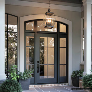 A modern front entryway with a black framed glass door, large windows, and potted plants is elegantly illuminated by the Urban Ambiance UQL1176 Colonial Outdoor Pendant Light from the Manchester Collection, featuring beveled glass and a Medieval Bronze finish, hanging strikingly above the entrance.