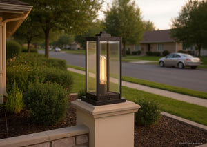 A sleek Estate Bronze outdoor pier light from the Quincy Collection by Urban Ambiance stands atop a square pillar near a suburban street lined with trees and houses. The UQL1355 Modern Farmhouse Outdoor Pier Light, measuring 16″ high by 7.5″ wide, features beveled glass for timeless elegance, adding distinctive charm and style to any setting.