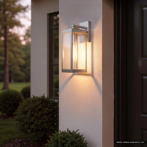 The UQL1361 Modern Farmhouse Outdoor Wall Light from the Quincy Collection by Urban Ambiance, featuring a sleek stainless steel finish and measuring 17 inches high by 6 inches wide, is illuminated on the exterior of a house near a dark door at dusk, with shrubs and trees in the background. This stylish wall-mounted fixture combines charm and functionality to enhance your entryway.