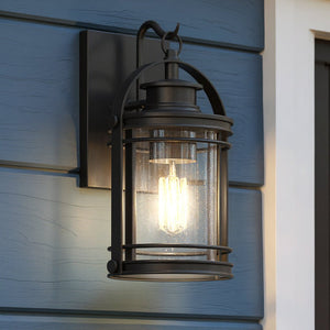 The UQL1430 Nautical Outdoor Wall Light from the Cannes Collection by Urban Ambiance, featuring an 11.5″ high by 6.75″ wide black silk finish and a clear glass shade, is illuminated and mounted on blue wood siding near a doorway, lending vintage-inspired lantern charm to your entryway.