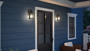 A black door with glass panels is centered on a blue house exterior, flanked by Urban Ambiance UQL1430 Nautical Outdoor Wall Lights from the Cannes Collection in a Black Silk finish. White trim, outdoor seating, and additional Black Silk aluminum accents are also visible.
