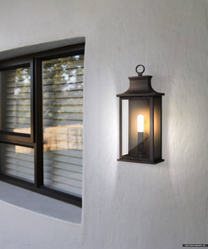 The Urban Ambiance UQL1721 Classic Outdoor Wall Sconce, 15.50''H, with an old bronze finish and marine-grade construction from the Halle Collection, is mounted next to a window with closed blinds on a white stucco wall.