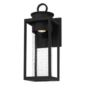 The Urban Ambiance UQL1780 Classic Outdoor Wall Sconce from the Nicosia Collection features marine-grade matte black aluminum and a cylindrical seeded glass shade, measuring 18.75''H x 7''W—ideal for adding style to any exterior space.