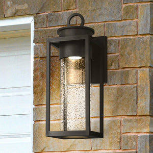 The Urban Ambiance UQL1780 Classic Outdoor Wall Sconce from the Nicosia Collection, featuring a matte black marine-grade finish, is mounted on a stone wall beside a white garage door.